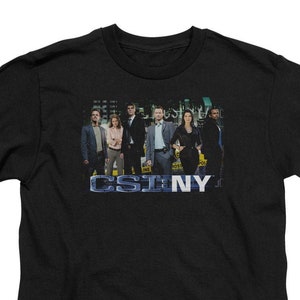 May include: Black t-shirt with a graphic of the cast of the TV show CSI: NY. The graphic features the characters standing in front of a city skyline. The text "CSI: NY" is printed in blue below the graphic.
