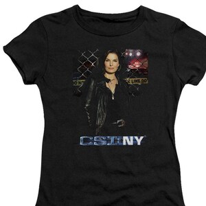 May include: A black t-shirt with a graphic of a woman in a leather jacket standing in front of a chain link fence. The text "CSI:NY" is printed in blue below the image.