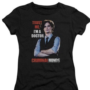 May include: Black t-shirt with a white graphic of a man wearing a vest and tie. The text "Trust Me, I'm a Doctor." is above the image and "Criminal Minds" is below the image.