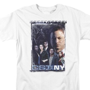 May include: White t-shirt with a graphic of the cast of CSI: NY. The graphic features a cityscape background with the cast members standing in front of it. The text "CSI: NY" is printed in blue at the bottom of the graphic.