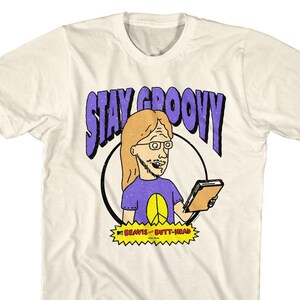 May include: A white t-shirt featuring a cartoon image of a character from the show Beavis and Butt-Head. The character is wearing a purple shirt with a peace sign and holding a book. The text "STAY GROOVY" is written in purple letters above the character. The text "BEAVIS and BUTT-HEAD" is written in red letters below the character.