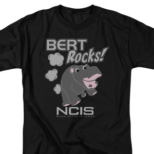 May include: Black t-shirt with a gray graphic of a cartoon hippopotamus with the text "BERT Rocks!" and "NCIS" below the graphic. The text "BASED ON THE TV SERIES" is below the "NCIS" logo.