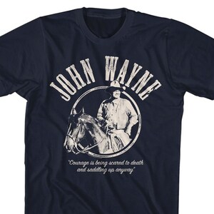 May include: A navy blue t-shirt featuring a white graphic of John Wayne riding a horse. The graphic is in a circle with the text "John Wayne" above it and the quote "Courage is being scared to death and saddling up anyway" below it.