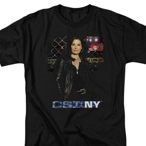 May include: Black t-shirt with a graphic of a woman in a leather jacket standing in front of a chain link fence. The text "CSI:NY" is printed in blue on the shirt.