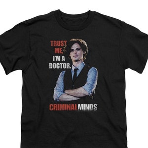 May include: Black t-shirt with a white graphic of a man in a suit and tie. The text "Trust me, I'm a doctor." is above the image and "Criminal Minds" is below the image.