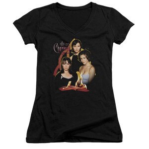 Puede incluir: Camiseta negra de cuello en V con un gr&aacute;fico de los tres personajes principales del programa de televisi&oacute;n "Charmed" de pie frente a un libro en llamas. El texto "Charmed" est&aacute; escrito en una fuente estilizada sobre los personajes.