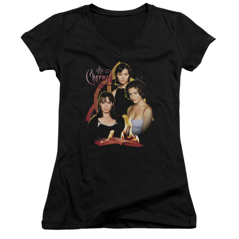 Puede incluir: Camiseta negra de cuello en V con un gr&aacute;fico de los tres personajes principales del programa de televisi&oacute;n "Charmed" de pie frente a un libro en llamas. El texto "Charmed" est&aacute; escrito en una fuente estilizada sobre los personajes.