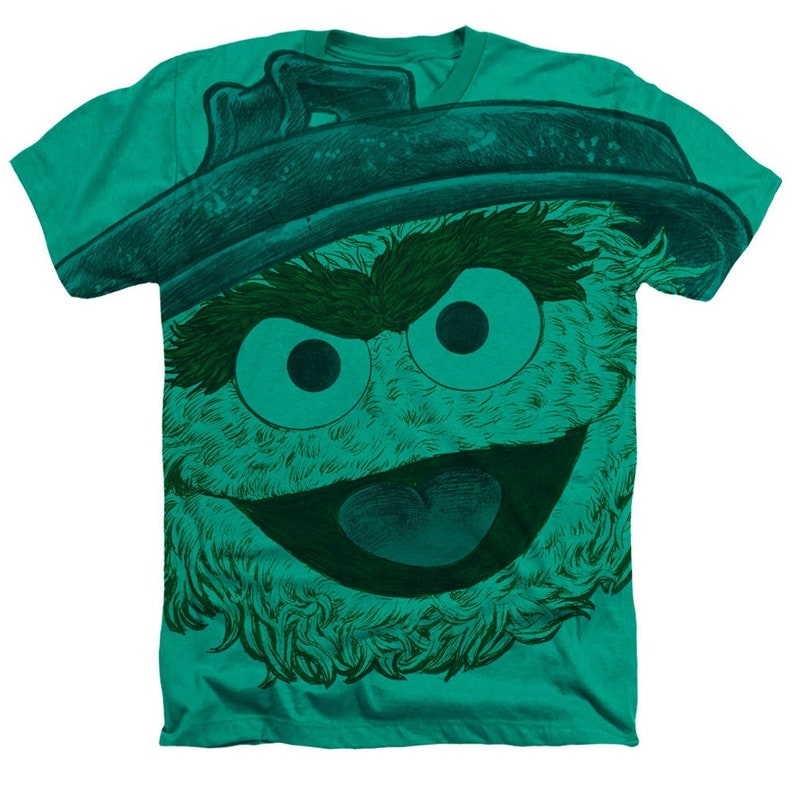 May include: A green t-shirt with a large, green, furry monster face graphic. The monster has a wide, toothy grin and a mischievous expression.