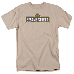 May include: Tan t-shirt featuring the Sesame Street sign. The sign is rectangular with a yellow border, black text, and the number 123 at the top. The shirt is made of a soft, neutral-colored fabric.