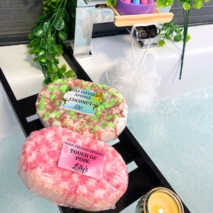 A shot of a bath, full of water and bubbles. Across the bath is a wooden tray, and on the tray are two Soap Infused Sponges: Coconut and Touch Of Pink. A small candle is lit to the side, showing a relaxing setting. Greenery surrounds the bathtub. The Coconut Sponge is white, green and brown, and the Touch Of Pink sponge is white and pink.