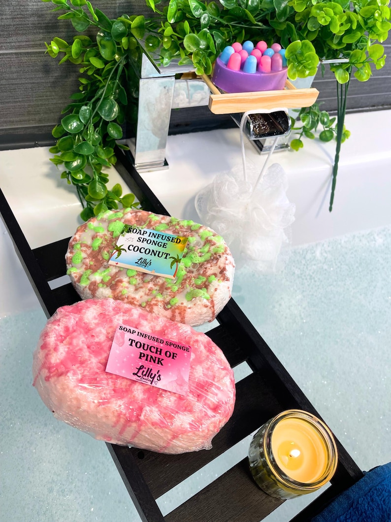 A shot of a bath, full of water and bubbles. Across the bath is a wooden tray, and on the tray are two Soap Infused Sponges: Coconut and Touch Of Pink. A small candle is lit to the side, showing a relaxing setting. Greenery surrounds the bathtub. The Coconut Sponge is white, green and brown, and the Touch Of Pink sponge is white and pink.