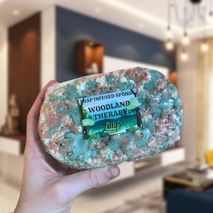 A Soap Infused Sponge being held up. The fragrance is Woodland Therapy, and the soap itself is decorated in colours reminiscent of the woods - primarily green, with brown wood-like soap also decorating the front, giving it a natural, eco design.
