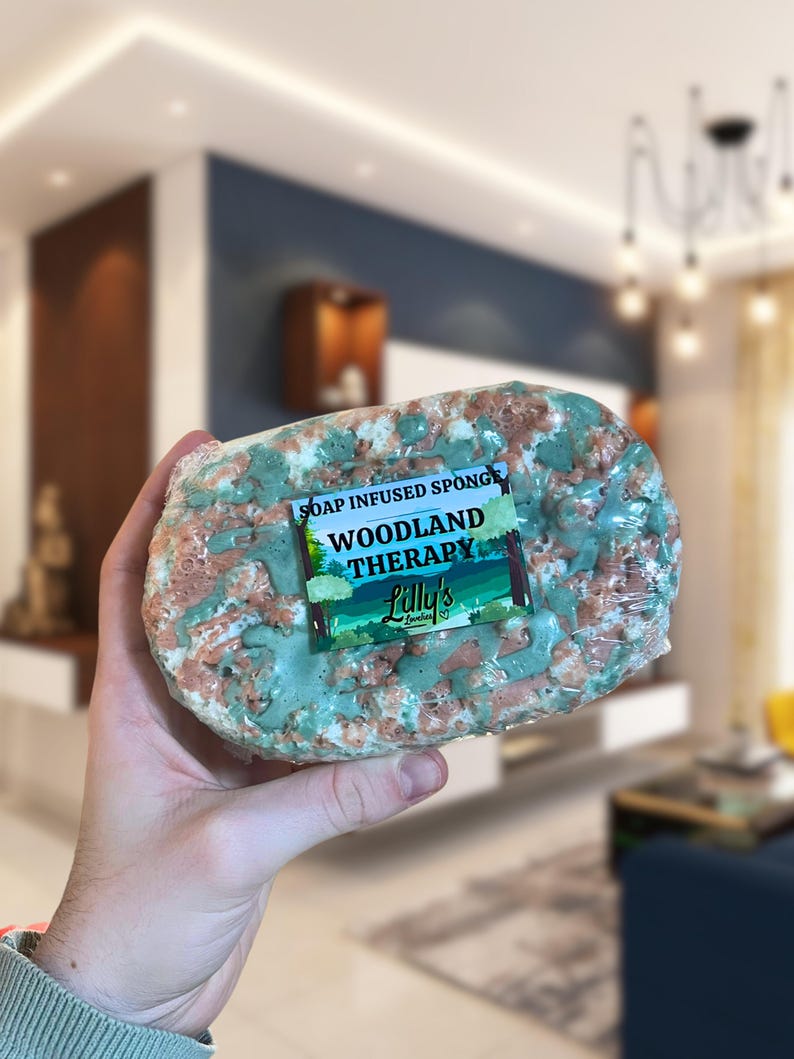 A Soap Infused Sponge being held up. The fragrance is Woodland Therapy, and the soap itself is decorated in colours reminiscent of the woods - primarily green, with brown wood-like soap also decorating the front, giving it a natural, eco design.