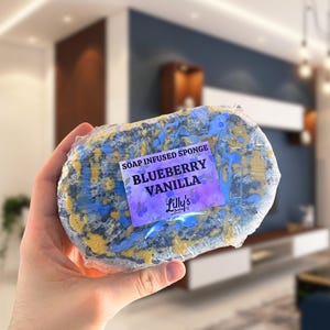 A shot of a Blueberry Vanilla Soap Infused Sponge being held up to show off the details. The sponge is purple and yellow. The sticker is placed on the front.