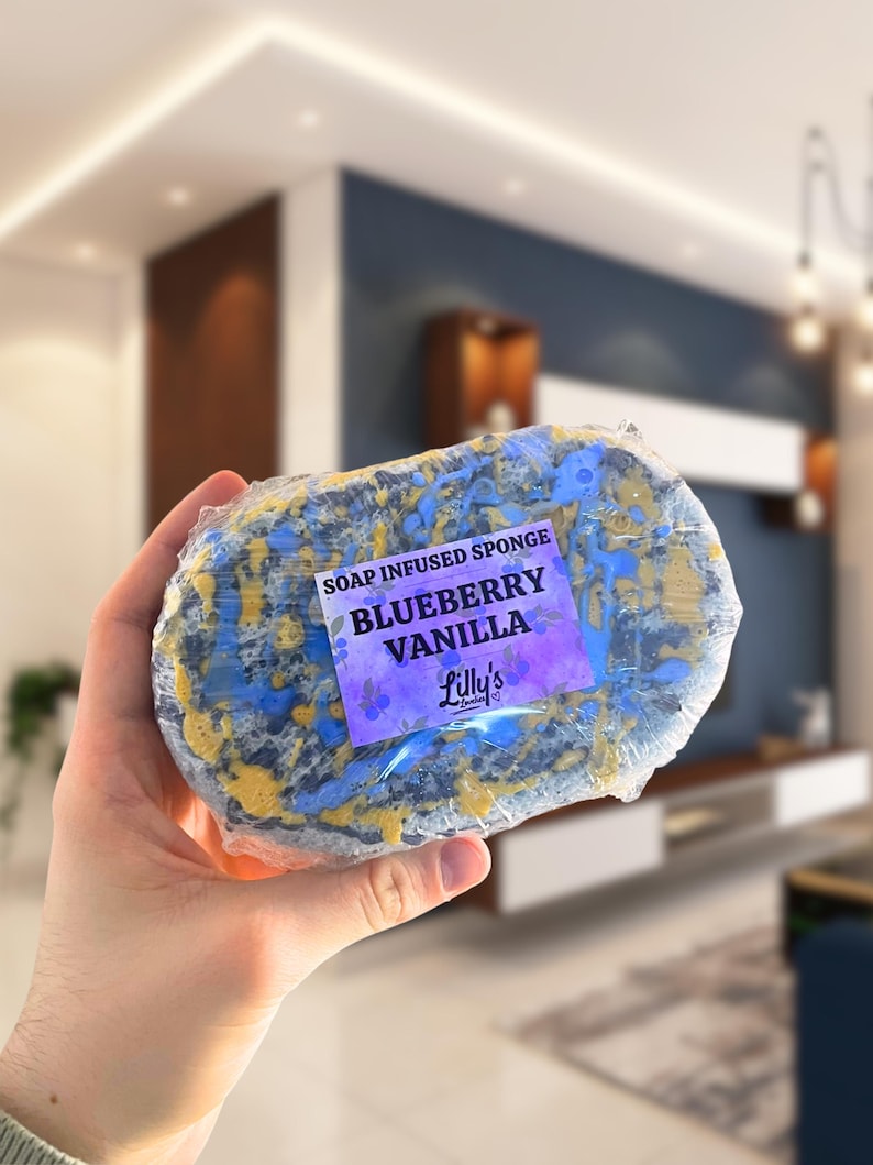 A shot of a Blueberry Vanilla Soap Infused Sponge being held up to show off the details. The sponge is purple and yellow. The sticker is placed on the front.