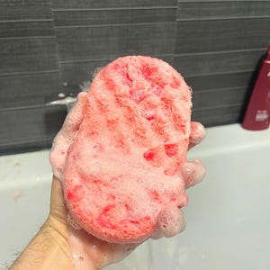 A wet Soap Infused Sponge, being held above bath water, covered in soapy suds.