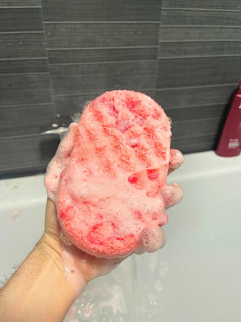 A wet Soap Infused Sponge, being held above bath water, covered in soapy suds.