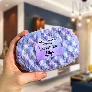A sponge held up to the camera, in focus against a blurred background. The Soap Infused Sponge is a Lavender fragrance, in deep purples of various shades. A sticker on the front displays the fragrance, and also shows the logo of the company, as well as a small butterfly.