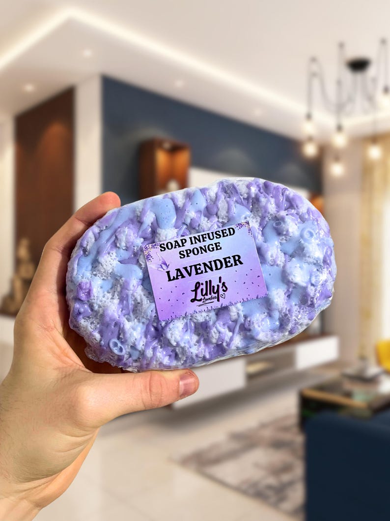 A sponge held up to the camera, in focus against a blurred background. The Soap Infused Sponge is a Lavender fragrance, in deep purples of various shades. A sticker on the front displays the fragrance, and also shows the logo of the company, as well as a small butterfly.
