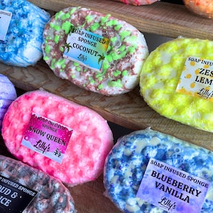 A large variety of different Soap Infused Sponges, displayed on a wooden shelf. They are all different in scent, with each decorated uniquely. Some of the sponges on display include: Coconut, Zesty Lemon, Blueberry Vanilla and Snow Queen. Each have soap topping them in different corresponding colours. For example, Zesty Lemon has yellow and white, while Blueberry Vanilla has blue, yellow and lilacs.