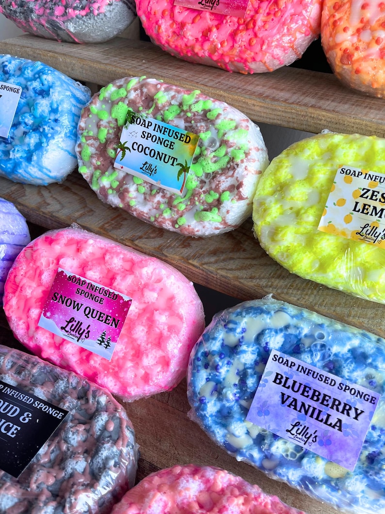 A large variety of different Soap Infused Sponges, displayed on a wooden shelf. They are all different in scent, with each decorated uniquely. Some of the sponges on display include: Coconut, Zesty Lemon, Blueberry Vanilla and Snow Queen. Each have soap topping them in different corresponding colours. For example, Zesty Lemon has yellow and white, while Blueberry Vanilla has blue, yellow and lilacs.