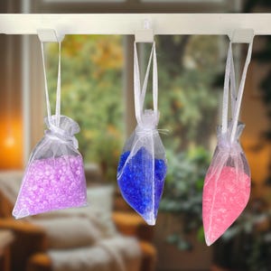 May include: Three sheer white organza drawstring pouches hanging from white hooks. The left pouch contains light purple beads, the centre pouch holds deep blue beads, and the right pouch is filled with bright pink beads. Each bag has a ribbon tie.