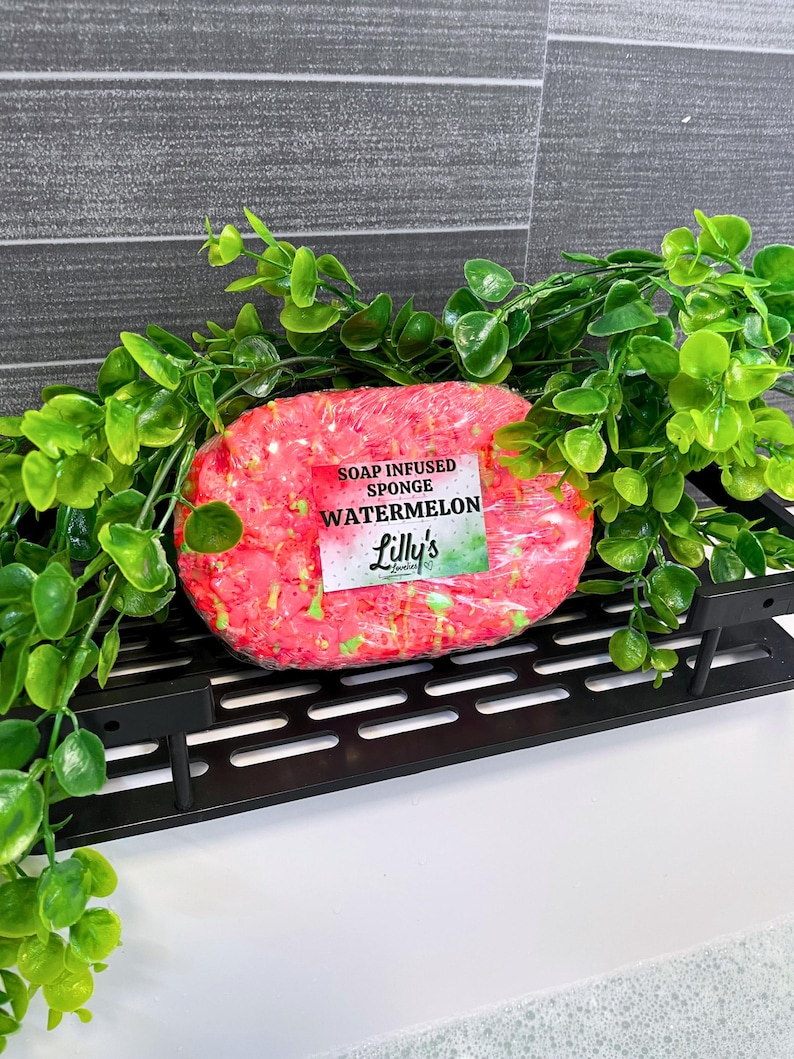A Watermelon Soap Infused Sponge, resting on a black tray on the side of a bubble-filled bath tub. The watermelon sponge is primarily red, topped off with green soap. It is surrounded by greenery and gives off natural vibes, reflecting the vegan properties of the Soap Infused Sponge itself.
