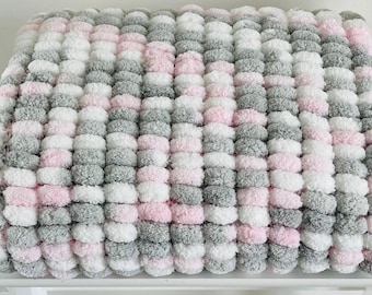 May include: A soft, plush blanket made of fluffy yarn in shades of pink, white, and gray. The blanket is made of a series of pom-poms, creating a unique and textured look.