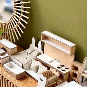 Puede incluir: Una casa de mu&ntilde;ecas de madera en miniatura con una cocina, un dormitorio y un ba&ntilde;o blancos. La casa de mu&ntilde;ecas incluye una cuna mecedora blanca, un sof&aacute; blanco, una ba&ntilde;era blanca y una mesa y sillas blancas. La casa de mu&ntilde;ecas est&aacute; hecha de madera y tiene un acabado de madera natural.