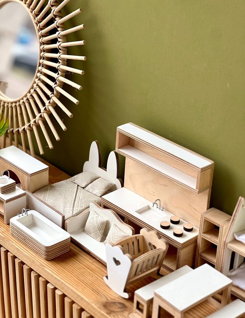 Puede incluir: Una casa de mu&ntilde;ecas de madera en miniatura con una cocina, un dormitorio y un ba&ntilde;o blancos. La casa de mu&ntilde;ecas incluye una cuna mecedora blanca, un sof&aacute; blanco, una ba&ntilde;era blanca y una mesa y sillas blancas. La casa de mu&ntilde;ecas est&aacute; hecha de madera y tiene un acabado de madera natural.