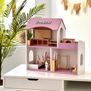 Puede incluir: Una casa de mu&ntilde;ecas de madera rosa y blanca con la palabra "Princess" en el techo. La casa de mu&ntilde;ecas tiene varias habitaciones, incluyendo un dormitorio, una cocina y un ba&ntilde;o. La casa de mu&ntilde;ecas est&aacute; sentada en una mesa blanca con un caj&oacute;n.