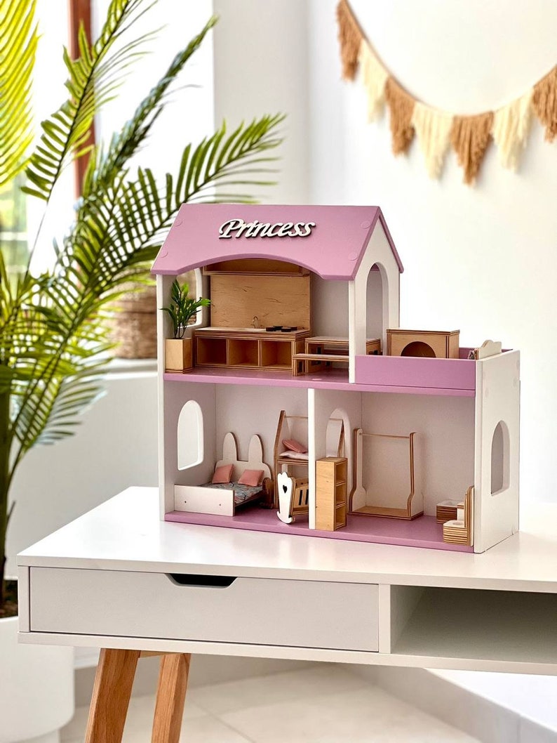 Puede incluir: Una casa de mu&ntilde;ecas de madera rosa y blanca con la palabra "Princess" en el techo. La casa de mu&ntilde;ecas tiene varias habitaciones, incluyendo un dormitorio, una cocina y un ba&ntilde;o. La casa de mu&ntilde;ecas est&aacute; sentada en una mesa blanca con un caj&oacute;n.