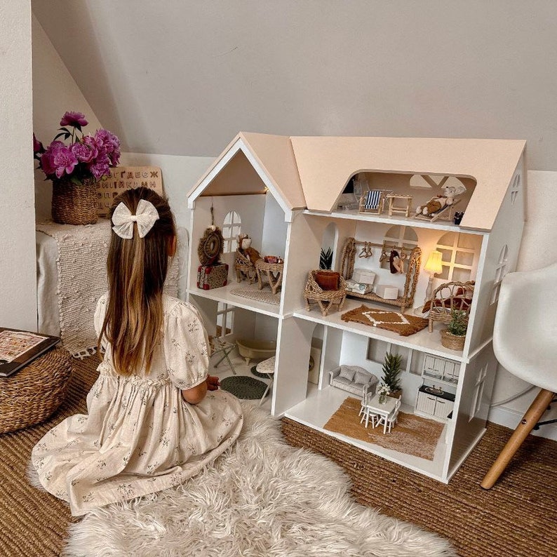 Op de afbeelding: Een groot poppenhuis met meerdere verdiepingen, een witte buitenkant en een lichtroze dak. Het poppenhuis is ingericht met miniatuurmeubels en decoraties. Een jong persoon zit op een zachte witte vloerkleed en kijkt naar het poppenhuis.