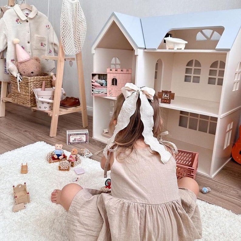 Op de afbeelding: Een kind in een beige jurk met een grote strik speelt met een poppenhuis op een wit tapijt. Het poppenhuis is wit met een lichtblauw dak. Miniatuurmeubels en speelgoed liggen verspreid.