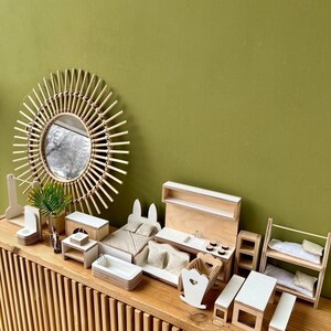 Puede incluir: Un conjunto de muebles de madera en miniatura para una casa de mu&ntilde;ecas, que incluye una cama, una cocina, una litera, una mecedora, una mesa y una mesita de noche. Los muebles est&aacute;n pintados de blanco y tienen un acabado de madera natural. Los muebles est&aacute;n dispuestos en un estante de madera con una pared verde en el fondo.