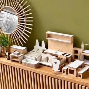 Puede incluir: Un conjunto de muebles de madera en miniatura para una casa de mu&ntilde;ecas, que incluye una cama, una cocina, una litera, una mesa y una mecedora. Los muebles est&aacute;n hechos de madera clara con detalles blancos.