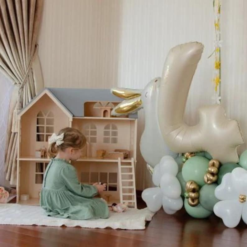 Op de afbeelding: Een houten poppenhuis met een grijs dak en een kleine ladder. Een jong meisje in een groene jurk zit op een wit tapijt en speelt met het poppenhuis. Een grote ballon met het cijfer 4 en andere ballonnen staan op de achtergrond.
