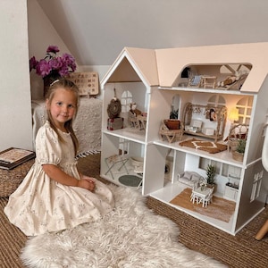 Op de afbeelding: Een jong meisje zit naast een groot poppenhuis met meerdere verdiepingen, met een witte buitenkant en een lichtroze dak. Het poppenhuis is ingericht met miniatuurmeubels en decoraties. Het meisje draagt een bloemenjurk en zit op een zachte witte vloerkleed.