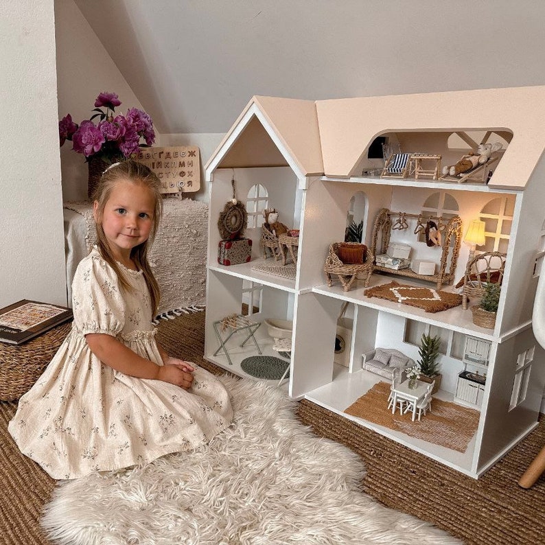 Op de afbeelding: Een jong meisje zit naast een groot poppenhuis met meerdere verdiepingen, met een witte buitenkant en een lichtroze dak. Het poppenhuis is ingericht met miniatuurmeubels en decoraties. Het meisje draagt een bloemenjurk en zit op een zachte witte vloerkleed.