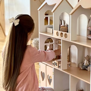 Può includere: Una casetta di bambole in legno bianca e beige con più stanze e una bambina che ci gioca. La casetta di bambole ha una porta e finestre a forma di cuore.