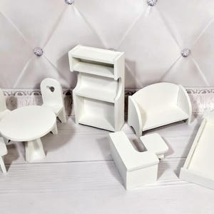 Op de afbeelding: Een verzameling miniatuur witte poppenhuismeubels, waaronder een tafel met stoelen, een boekenplank, een bank, een bed en een bureau. De stoelen hebben hartvormige uitsparingen. De meubels zijn op een wit oppervlak geplaatst.