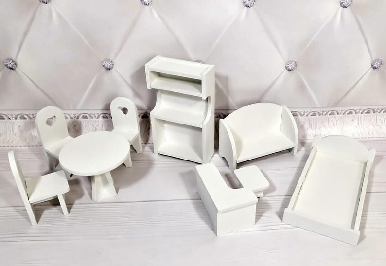 Op de afbeelding: Een verzameling miniatuur witte poppenhuismeubels, waaronder een tafel met stoelen, een boekenplank, een bank, een bed en een bureau. De stoelen hebben hartvormige uitsparingen. De meubels zijn op een wit oppervlak geplaatst.