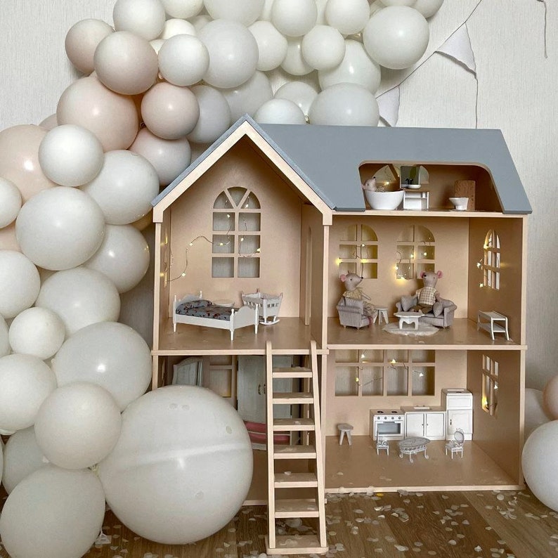 Op de afbeelding: Een houten poppenhuis met meerdere verdiepingen, een grijs dak en een lichte houten buitenkant. Het poppenhuis is ingericht met miniatuurmeubels en heeft een ladder. De achtergrond toont witte en roze ballonnen.