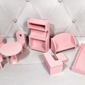 Op de afbeelding: Een verzameling miniatuur roze houten poppenhuismeubels, waaronder een tafel met twee stoelen, een boekenplank, een bank, een bureau en een bedframe. De stoelen hebben hartvormige uitsparingen.