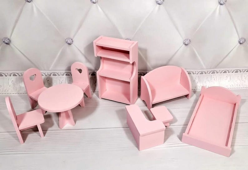 Op de afbeelding: Een verzameling miniatuur roze houten poppenhuismeubels, waaronder een tafel met twee stoelen, een boekenplank, een bank, een bureau en een bedframe. De stoelen hebben hartvormige uitsparingen.
