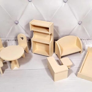 Op de afbeelding: Een verzameling onafgewerkte houten poppenhuismeubels, waaronder een tafel met stoelen, een boekenplank, een bank, een bed en een bijzettafel. De stoelen hebben hartvormige uitsparingen. De meubels zijn lichtbeige van kleur.