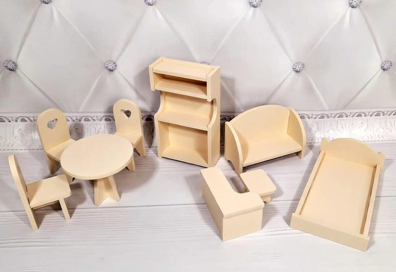 Op de afbeelding: Een verzameling onafgewerkte houten poppenhuismeubels, waaronder een tafel met stoelen, een boekenplank, een bank, een bed en een bijzettafel. De stoelen hebben hartvormige uitsparingen. De meubels zijn lichtbeige van kleur.