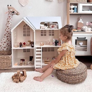 Op de afbeelding: Een kind zit in de buurt van een poppenhuis met miniatuurmeubels en accessoires. Het poppenhuis is wit met een lichtblauw dak. Een giraffenknuffel en een rieten mand staan ook in de kamer. Houten blokken met cijfers liggen op de vloer.