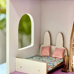 Puede incluir: Una cama de madera blanca para casa de mu&ntilde;ecas con un edred&oacute;n estampado floral y dos almohadas rosas. La cama est&aacute; en una habitaci&oacute;n de casa de mu&ntilde;ecas con una pared blanca y un recorte de ventana.