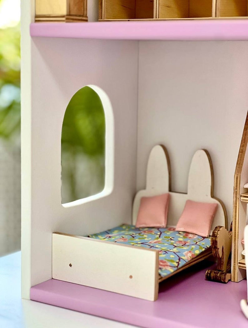 Puede incluir: Una cama de madera blanca para casa de mu&ntilde;ecas con un edred&oacute;n estampado floral y dos almohadas rosas. La cama est&aacute; en una habitaci&oacute;n de casa de mu&ntilde;ecas con una pared blanca y un recorte de ventana.